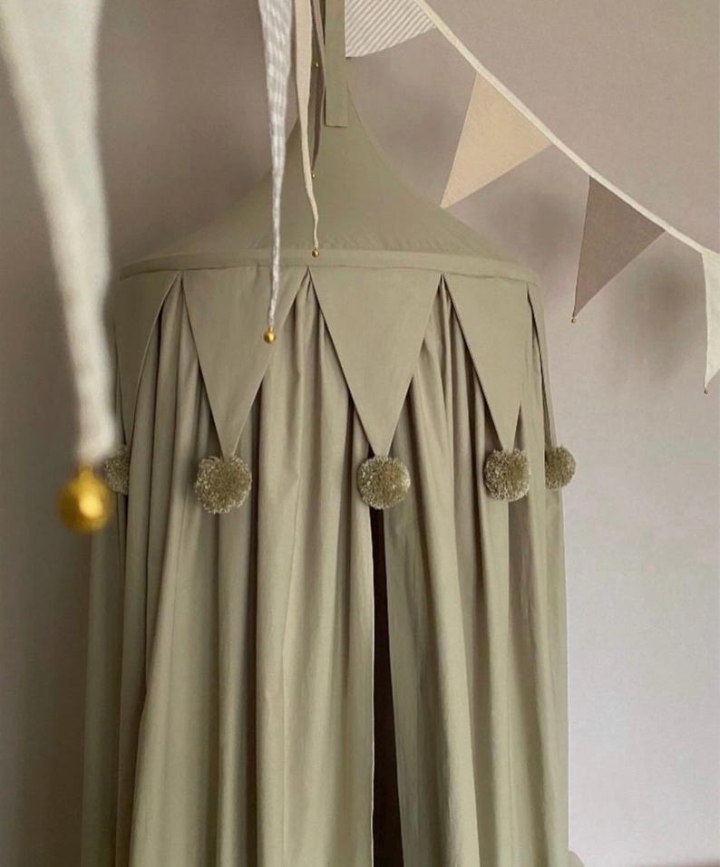 Pu&ograve; includere: Un baldacchino verde salvia con finiture triangolari e pompon dorati. Il baldacchino &egrave; sospeso al soffitto e presenta un design drappeggiato. Una ghirlanda di gagliardetti dai toni neutri &egrave; appesa nelle vicinanze, aggiungendo un tocco decorativo.