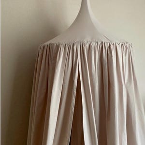 Pu&ograve; includere: Un letto a baldacchino beige con una parte superiore conica e tessuto arricciato. Il tessuto scende, creando un effetto morbido e fluido. Il baldacchino &egrave; sospeso al soffitto, offrendo uno spazio accogliente e chiuso.