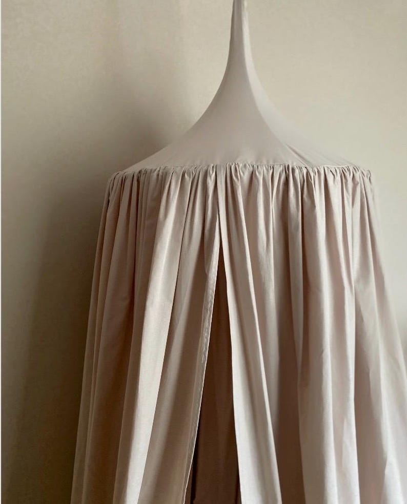Pu&ograve; includere: Un letto a baldacchino beige con una parte superiore conica e tessuto arricciato. Il tessuto scende, creando un effetto morbido e fluido. Il baldacchino &egrave; sospeso al soffitto, offrendo uno spazio accogliente e chiuso.