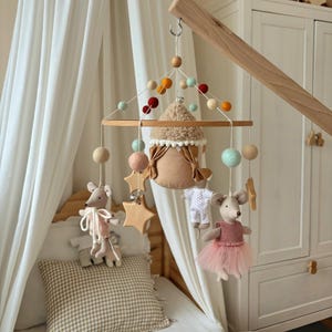 May include: A handmade baby mobile with a wooden frame, featuring felt balls in various colors, star-shaped wooden cutouts, and plush mouse figures. The mobile hangs above a crib with a canopy and a checkered pillow.