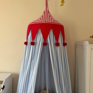 May include: A hanging fabric canopy featuring a red and white striped conical top with a red scalloped border adorned with small red pom-poms. The main body of the canopy consists of light blue and white vertical striped fabric panels that drape down.