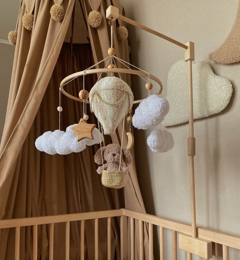 May include: A wooden baby mobile with a brown teddy bear in a basket, a white hot air balloon, white clouds, a wooden star, and a crescent moon. The mobile is hanging from a wooden stand.