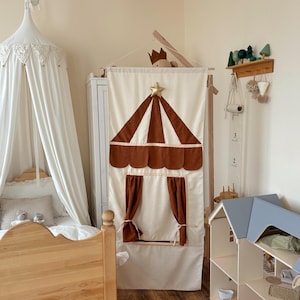 May include: A cream-colored fabric wall hanging featuring a brown circus tent design with a gold star at the top and tie-back curtains. The piece is suspended from a wooden rod and displays a scalloped awning detail above the window opening.