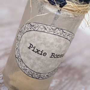May include: A clear glass bottle with a label that reads "Pixie Bones". The bottle is tied with twine and has a black wax seal.