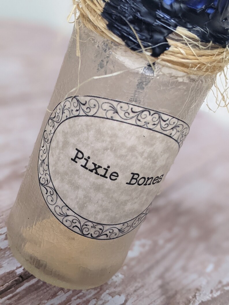 May include: A clear glass bottle with a label that reads "Pixie Bones". The bottle is tied with twine and has a black wax seal.