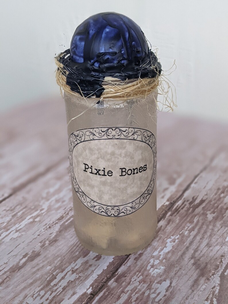 May include: A glass jar with a black and blue wax seal and a label that reads "Pixie Bones". The jar is filled with a clear liquid and has a straw-like object inside.