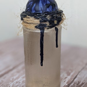 May include: A clear glass bottle with a blue and black orb on top. The orb is surrounded by a woven straw nest. Black paint drips down the side of the bottle.