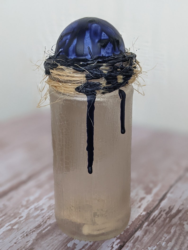 May include: A clear glass bottle with a blue and black orb on top. The orb is surrounded by a woven straw nest. Black paint drips down the side of the bottle.