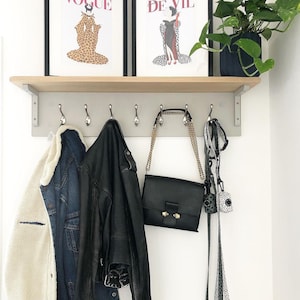 Puede incluir: Un perchero de madera con ganchos, con dos impresiones enmarcadas de portadas de las revistas Vogue y De Vil. Un bolso de cuero negro, una chaqueta vaquera y una chaqueta de sherpa blanca y negra cuelgan de los ganchos. Una planta en maceta est&aacute; en la repisa sobre el perchero.