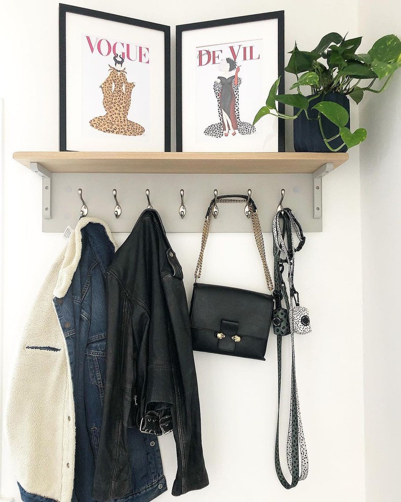 Puede incluir: Un perchero de madera con ganchos, con dos impresiones enmarcadas de portadas de las revistas Vogue y De Vil. Un bolso de cuero negro, una chaqueta vaquera y una chaqueta de sherpa blanca y negra cuelgan de los ganchos. Una planta en maceta est&aacute; en la repisa sobre el perchero.