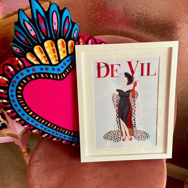 Puede incluir: Una impresi&oacute;n enmarcada en blanco con una ilustraci&oacute;n en blanco y negro de Cruella de Vil con un abrigo de estampado d&aacute;lmata y un vestido negro. El texto "Cruella de Vil" est&aacute; en letras rojas en la impresi&oacute;n.