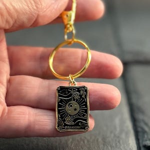 May include: A gold-toned metal keyring featuring a rectangular black enamel charm. The charm displays a sun and moon design with celestial rays and hands, with the text THE MAGICIAN printed at the bottom. The keyring includes a circular ring.