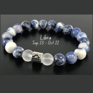 May include: A beaded stretch bracelet featuring round blue and white mottled stone beads, two clear frosted beads, and a central silver-toned metal charm embossed with the Libra zodiac symbol. The text Libra Sep 23 - Oct 22 is visible.