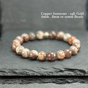 May include: A beaded bracelet made of copper sunstone and 14K gold accents. The text Copper Sunstone - 14K Gold 6mm, 8mm or 10mm Beads is visible. The bracelet consists of round, polished, brownish-orange beads with a reflective, shimmering surface.