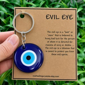 May include: A circular blue glass evil eye charm attached to a silver metal keyring, mounted on a rectangular brown card. The card features the text EVIL EYE and a paragraph explaining the talisman, with the URL ccshealingcrystals.etsy.com.