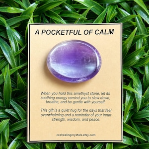 May include: A smooth, oval-shaped purple amethyst stone centred on a rectangular tan card. The card features the text A POCKETFUL OF CALM, followed by a message about holding the stone, and the website address ccshealingcrystals.etsy.com.