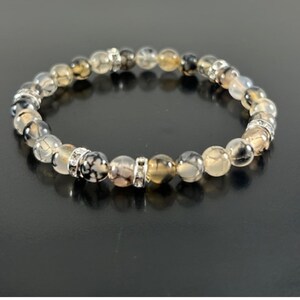 May include: A beaded bracelet featuring a series of round, marbled beads in shades of gray, brown, and cream. The bracelet includes silver-toned spacers with clear, sparkling accents. The bracelet is displayed on a dark surface.