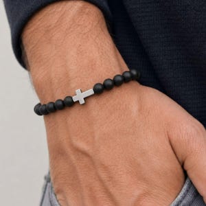 May include: A stretch bracelet featuring matte black spherical beads and a single silver-toned metal cross charm positioned centrally. The beads are uniform in size and arranged in a continuous circular pattern around the wrist.