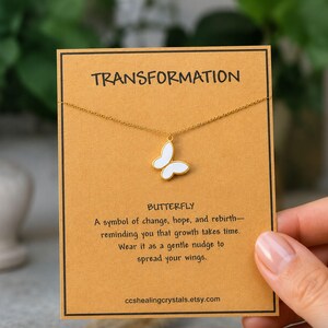 May include: A gold-tone chain necklace with a white butterfly pendant displayed on a tan card. The card features the text TRANSFORMATION, BUTTERFLY, and a paragraph about growth, along with the website ccshealingcrystals.etsy.com at the bottom.
