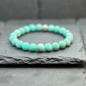 May include: A stretch bracelet composed of round, smooth, light turquoise-green amazonite beads. The beads are arranged in a circular pattern, featuring a single metallic silver-toned bead positioned at the top centre of the circular strand.