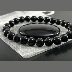 May include: A circular bracelet composed of smooth, polished black spherical beads arranged in a continuous loop. The beads have a reflective, glossy surface that catches the light, creating small white highlights on each individual bead.