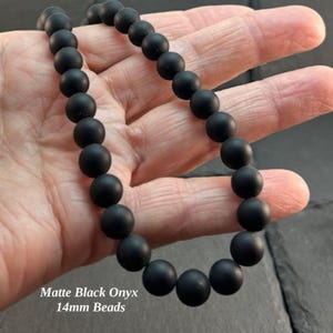 May include: A necklace featuring matte black onyx beads, each measuring 14mm in diameter. The beads are round, uniform in size, and strung together to form a continuous strand of dark, non-reflective stones that display a smooth, matte finish.
