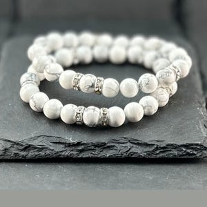 May include: Two white and gray beaded bracelets with silver accents. The bracelets are made of round, marble-like beads and are displayed on a dark gray surface. The bracelets are a stylish accessory.