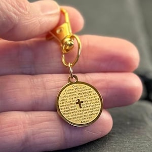May include: A gold-coloured circular metal charm attached to a lobster claw clasp. The charm features the Lord's Prayer engraved in black text on its surface, with a small cross symbol positioned in the centre of the prayer text.