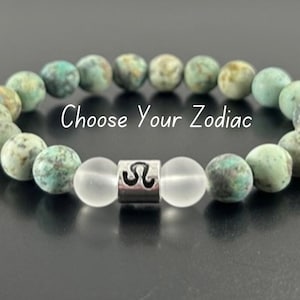 May include: A turquoise and white beaded bracelet with a silver charm featuring the astrological symbol for Leo. The bracelet has the text "Choose Your Zodiac" printed on it.