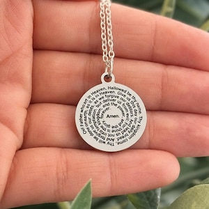 May include: A circular silver-coloured pendant necklace featuring the Lord's Prayer engraved in black text in a spiral pattern. The pendant is attached to a delicate silver-coloured chain link necklace, with the text ending in the word Amen at the centre.