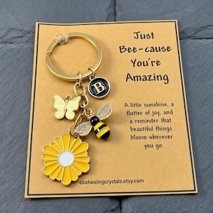 May include: A gold-toned metal keychain featuring a yellow daisy charm, a yellow butterfly charm, a bee charm with clear crystals, and a black circular charm with the letter B. The keychain is attached to a tan card with the text Just Bee-cause You're Amazing.