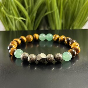 May include: A beaded bracelet featuring alternating brown and gold tiger eye beads, smooth light green aventurine spheres, and metallic pyrite beads. The center of the bracelet showcases a single faceted, geometric pyrite stone bead.
