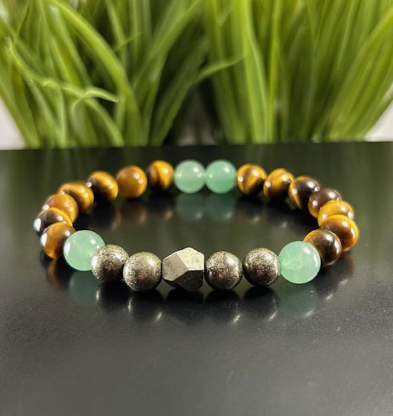 May include: A beaded bracelet featuring alternating brown and gold tiger eye beads, smooth light green aventurine spheres, and metallic pyrite beads. The center of the bracelet showcases a single faceted, geometric pyrite stone bead.