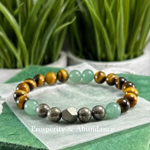 May include: A beaded bracelet featuring alternating brown and gold tiger eye beads, smooth light green jade-like beads, and metallic pyrite spheres, with a central faceted pyrite bead and the text Prosperity & Abundance printed below.
