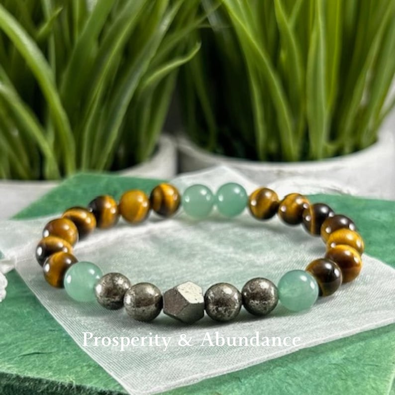 May include: A beaded bracelet featuring alternating brown and gold tiger eye beads, smooth light green jade-like beads, and metallic pyrite spheres, with a central faceted pyrite bead and the text Prosperity & Abundance printed below.