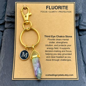 May include: A gold-toned keyring featuring a hexagonal fluorite crystal point and a small circular charm with the letter M in white on a black background. The keyring is attached to a tan display card labelled FLUORITE with descriptive text.