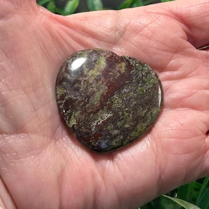 May include: A polished heart-shaped stone featuring a mottled pattern of deep burgundy, dark brown, and olive green colors. The surface is smooth and reflective, showcasing natural mineral variations throughout the stone's surface.