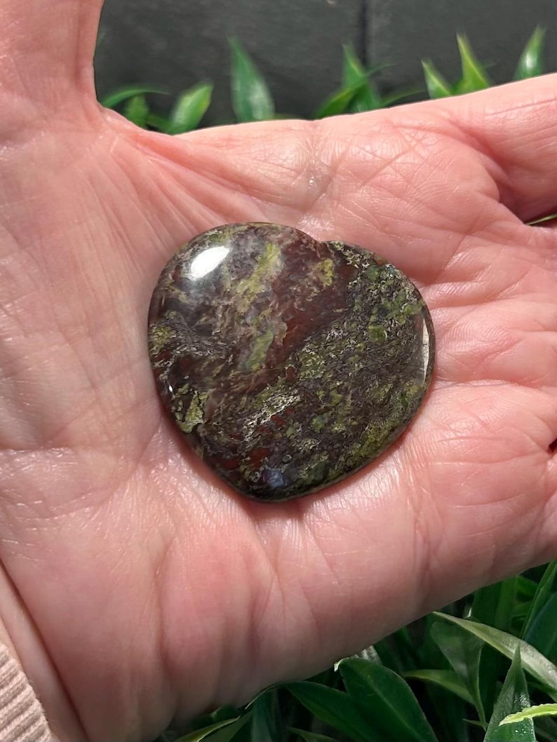 May include: A polished heart-shaped stone featuring a mottled pattern of deep burgundy, dark brown, and olive green colors. The surface is smooth and reflective, showcasing natural mineral variations throughout the stone's surface.