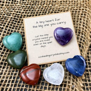 May include: A square beige card with the text A tiny heart for the big one you carry. Let this little crystal remind you you are loved. even on the quiet days. ccshealingcrystals.etsy.com. Six polished heart-shaped crystals in purple, green, dark green, red, clear, and blue.