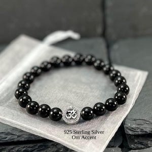 May include: A stretch bracelet composed of smooth, round, polished black beads. The bracelet features a single circular 925 sterling silver accent bead engraved with an Om symbol and a Greek key border pattern. The text 925 Sterling Silver Om Accent is visible.