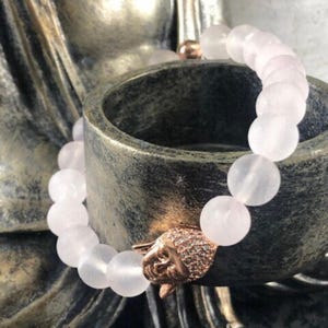 May include: A stretch bracelet composed of smooth, pale pink round beads. The bracelet features a single rose gold-toned charm shaped like a Buddha head, which is encrusted with small, clear sparkling stones on the top portion of the head.