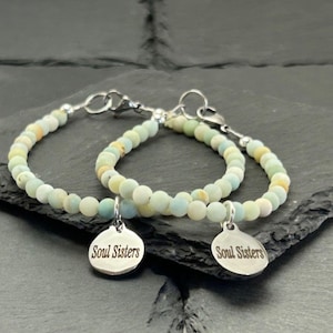 May include: Two beaded bracelets featuring light blue and cream-coloured matte stones. Each bracelet includes a round silver-toned metal charm engraved with the text Soul Sisters. The bracelets are secured with silver-toned lobster claw clasps.