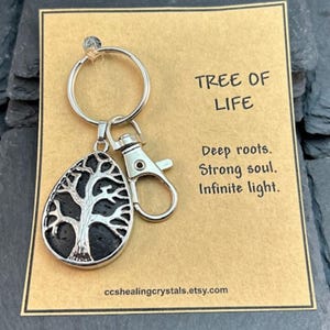 May include: A silver keychain with a tree of life pendant. The pendant has a black background and a silver tree design. The card behind the keychain reads "TREE OF LIFE" and "Deep roots. Strong soul. Infinite light."