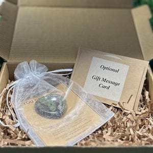 May include: A brown cardboard gift box filled with shredded paper, containing a small green stone heart inside a white organza drawstring pouch, and a tan card labeled Optional Gift Message Card with printed text.