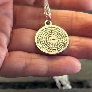 Peut inclure: Un pendentif circulaire de couleur argent&eacute;e avec la pri&egrave;re du Notre P&egrave;re grav&eacute;e en noir. Le texte commence par Our Father who art in Heaven et se termine par Amen au centre. Le pendentif est suspendu &agrave; une fine cha&icirc;ne de couleur argent&eacute;e.