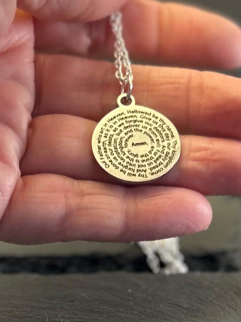 Peut inclure: Un pendentif circulaire de couleur argent&eacute;e avec la pri&egrave;re du Notre P&egrave;re grav&eacute;e en noir. Le texte commence par Our Father who art in Heaven et se termine par Amen au centre. Le pendentif est suspendu &agrave; une fine cha&icirc;ne de couleur argent&eacute;e.