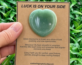 May include: A smooth, heart-shaped green aventurine stone attached to a rectangular tan card. The card features the text LUCK IS ON YOUR SIDE at the top, followed by descriptive text about the stone and the website ccshealingcrystals.etsy.com.