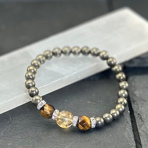 May include: A beaded bracelet featuring metallic gray beads and accent stones. The bracelet includes two brown tiger's eye stones and a yellow citrine stone, each separated by silver-colored spacers with clear crystals. The bracelet is displayed on a white surface.
