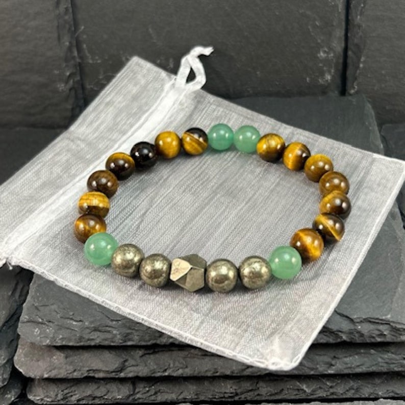 May include: A beaded bracelet featuring a sequence of round brown tiger's eye beads, smooth green aventurine beads, and metallic-toned pyrite beads, including one central faceted pyrite bead, arranged in a circular pattern on a light gray fabric pouch.