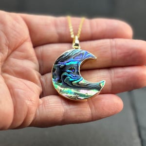 May include: A gold necklace with a crescent moon pendant. The pendant is inlaid with iridescent abalone shell, displaying blues, greens, and purples. This delicate necklace is a piece of jewellery.